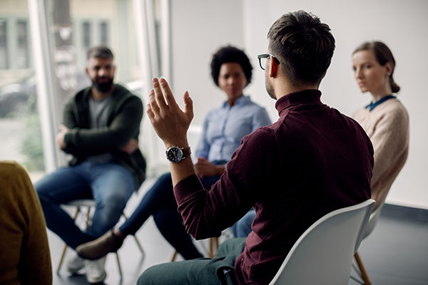 Rear view of man raising his hand to ask a question while attending group therapy.